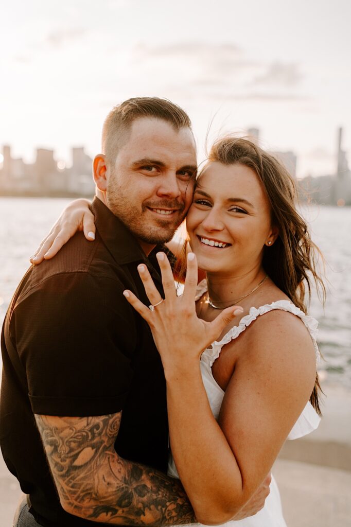 A couple smile at the camera and embrace as the woman shows off her hand which has an engagement ring on it, behind them is the Chicago skyline and Lake Michigan