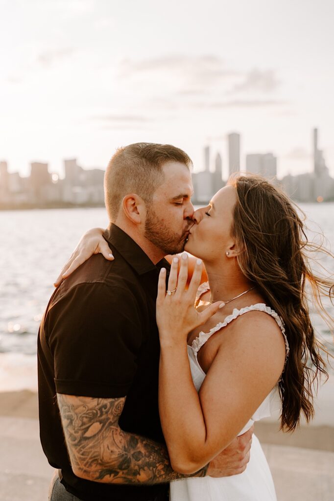 A couple standing in front of Lake Michigan and the Chicago skyline kiss one another while the woman shows off her engagement ring on her hand