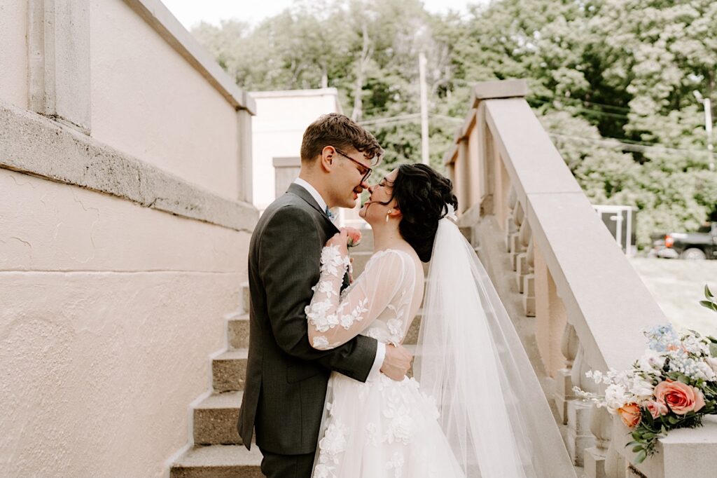 Outside their wedding venue the Fowler House Mansion, a bride and groom embrace and are about to kiss while walking up the stone stairway outdoors
