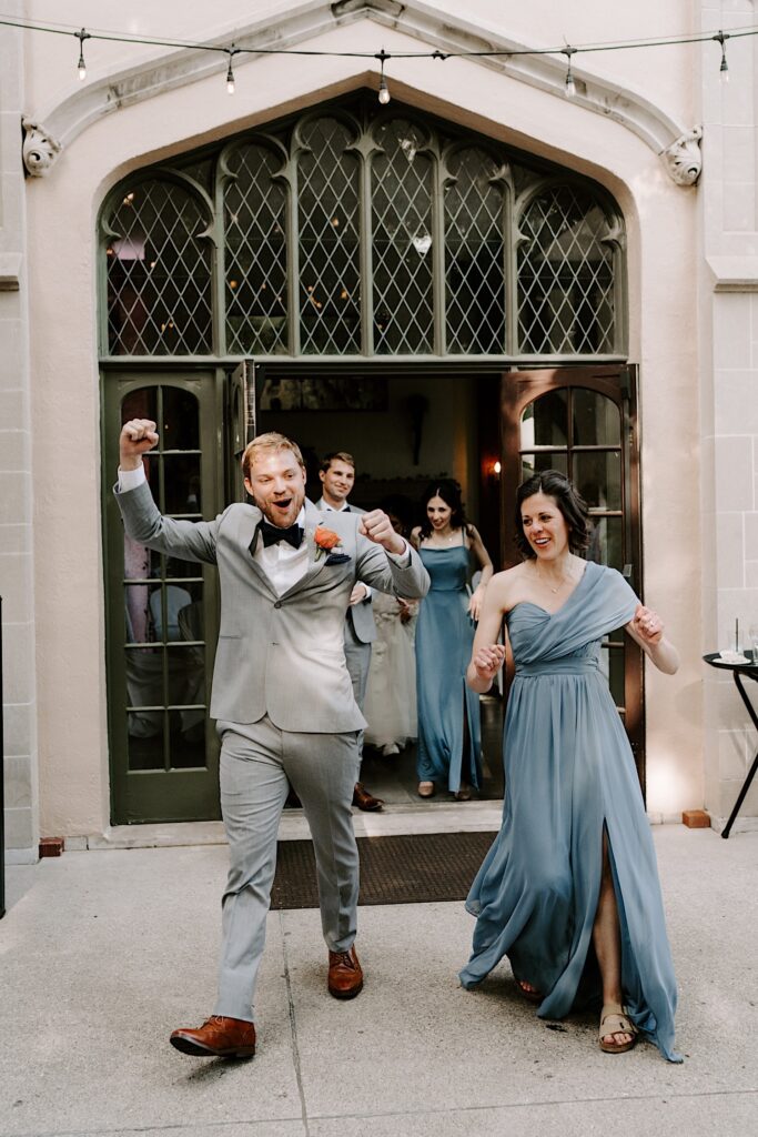 A groomsman and a bridesmaid walk into a wedding reception together and celebrate with other members of the wedding parties behind them