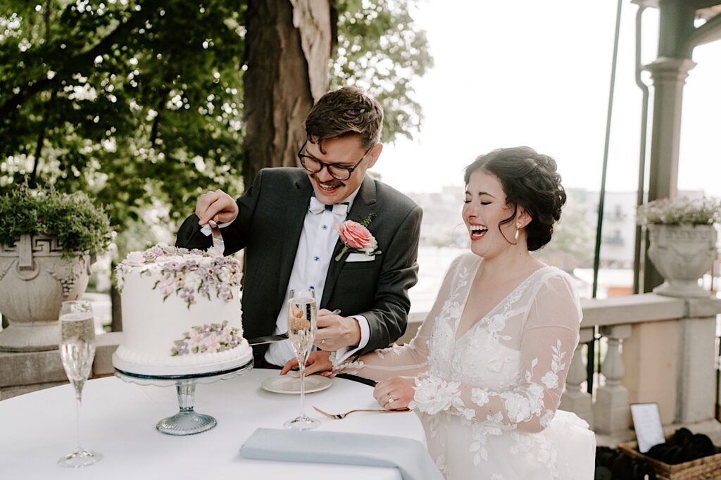A bride and groom smile while cutting their wedding cake on an outdoor patio at their wedding reception at the Fowler House Mansion