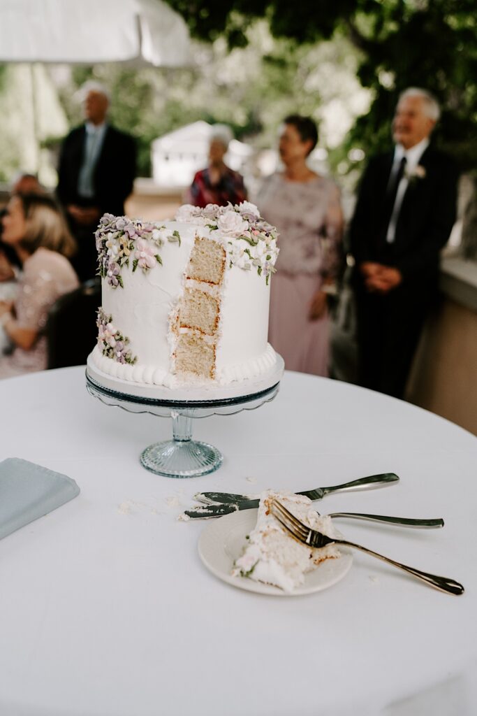 A wedding cake on a table sits on a platter with a piece cut out of it that sits on a plate with a fork nearby
