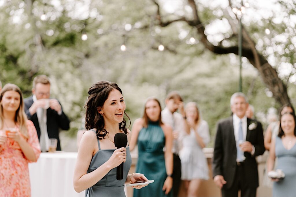 A bridesmaid stands with a microphone and gives a speech during an outdoor wedding reception at the Fowler House Mansion