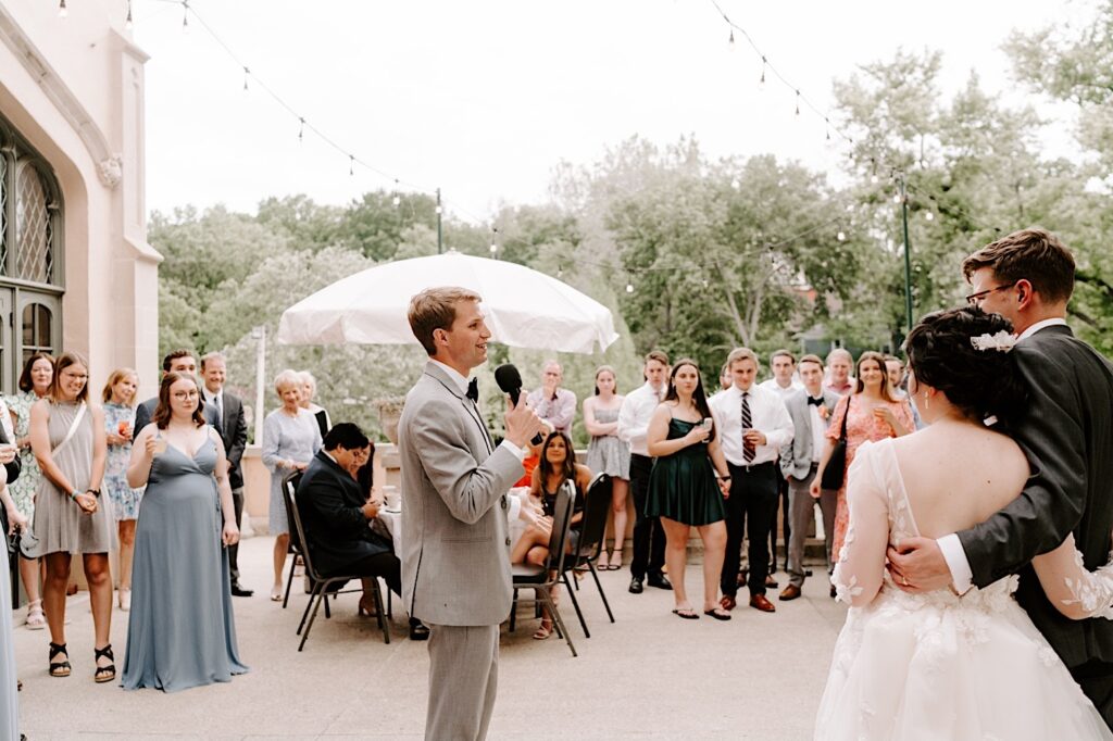 A bride and groom stand next to one another and hold each other with their backs to the camera looking at a groomsmen who's giving a speech during their outdoor wedding reception at the Fowler House Mansion