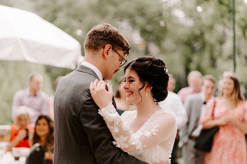 A bride smiles while looking at the groom as they embrace during their first dance at their outdoor wedding reception at the Fowler House Mansion