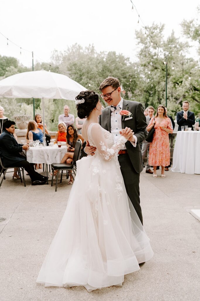 A bride and groom embrace as they dance with one another during their outdoor wedding reception surrounded by their guests