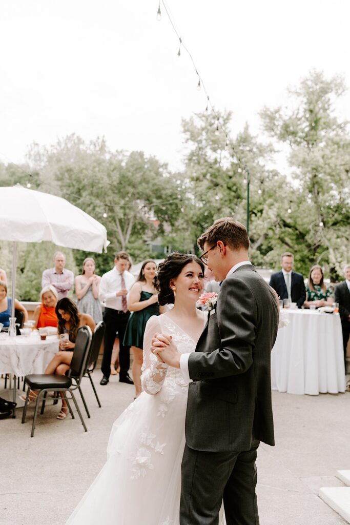 A bride and groom embrace and smile as they dance with one another during their outdoor wedding reception surrounded by their guests