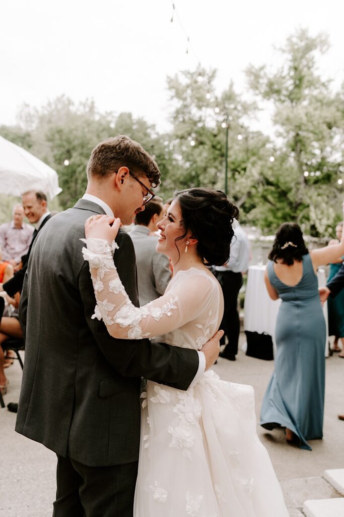 A bride and groom smile at one another as they dance with the guests of their wedding during their outdoor wedding reception