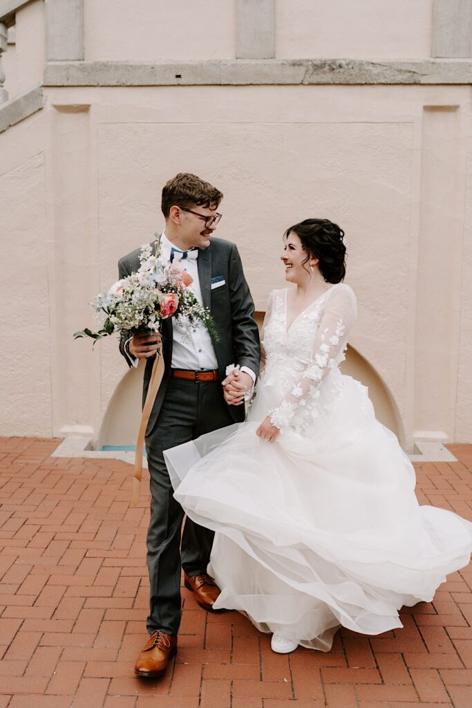 A bride and groom walk while holding hands and smiling at one another on a brick patio
