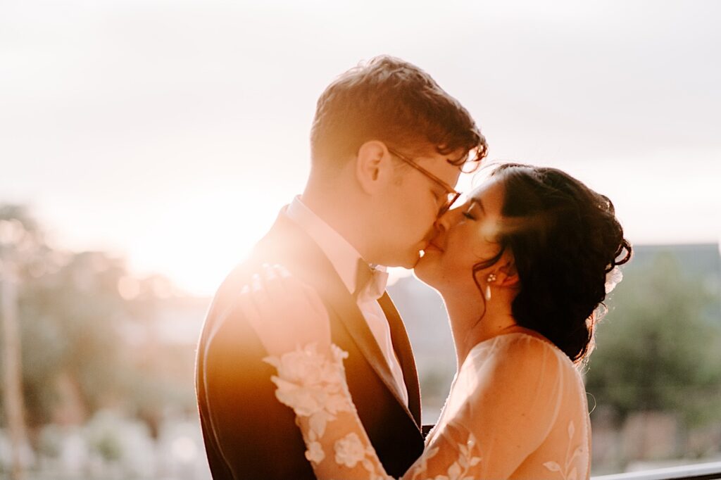 During their outdoor wedding reception at the Fowler House Mansion, a bride and groom kiss one another with the sun setting behind them