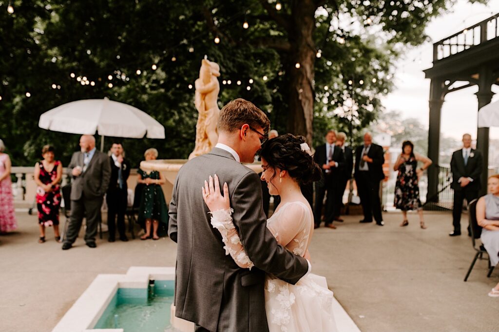 A bride and groom facing away from the camera stand next to a fountain and embrace during their outdoor wedding reception at the Fowler House Mansion