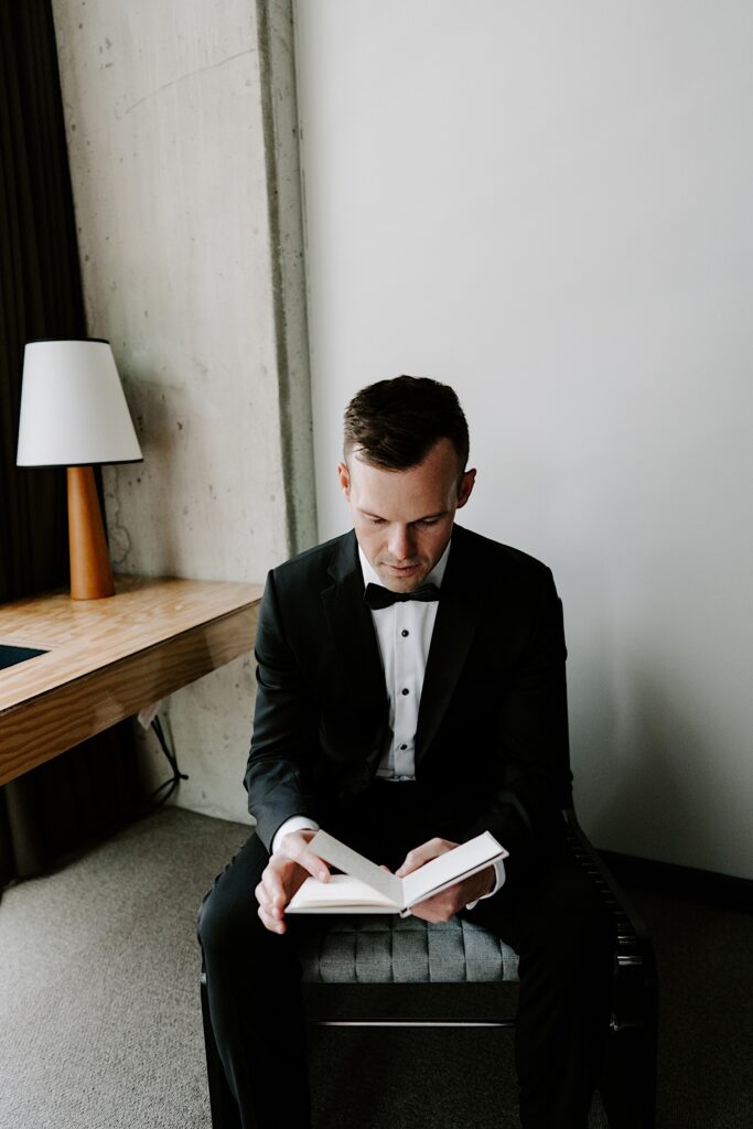 A groom in a wedding tux sits on a chair next to a desk and reads over his vows