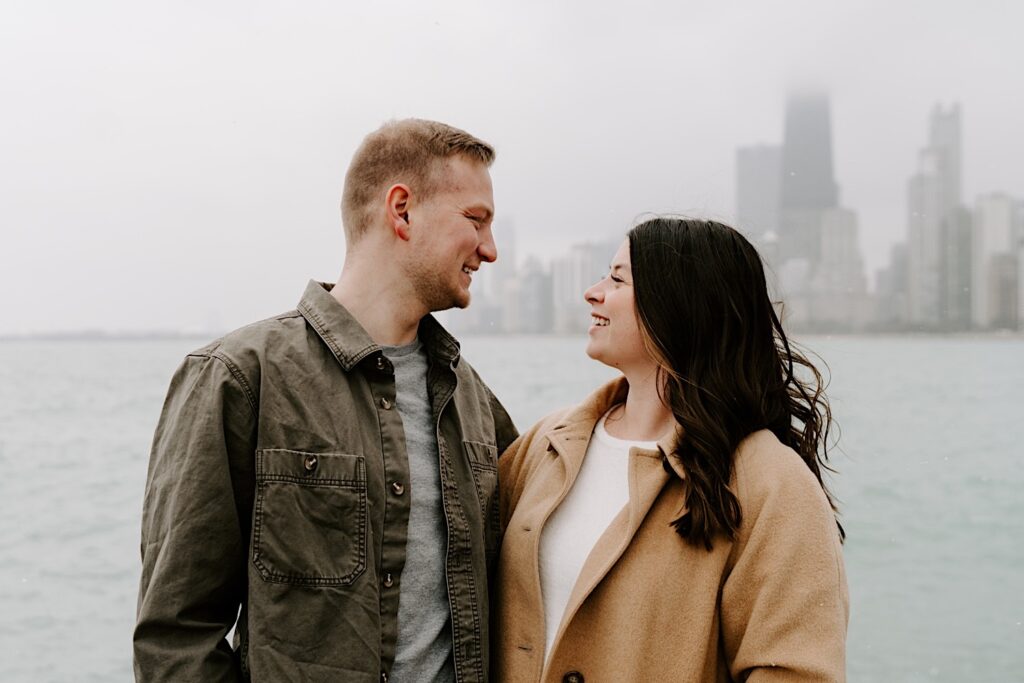 A couple smile at one another while standing next to each other in with Lake Michigan and the Chicago skyline behind them during their Fall Engagement Session at North Avenue Beach