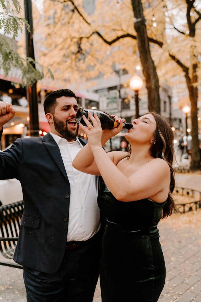 A woman drinks from a bottle of champagne as a man next to her cheers her on during their fall engagement session in Chicago