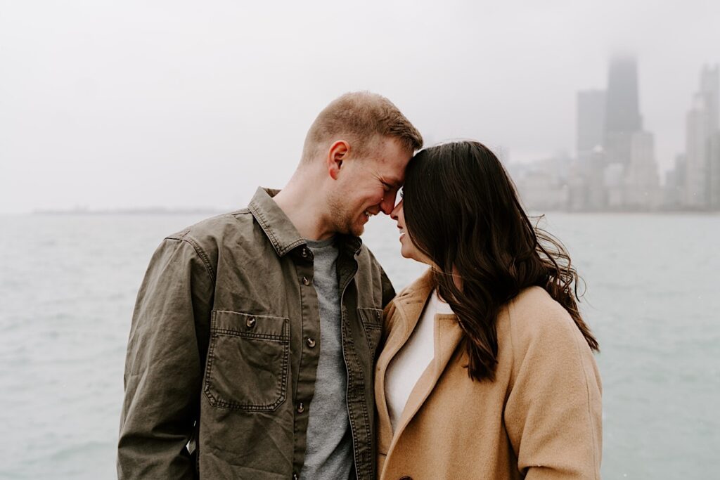 A couple smile at one another while touching noses in front of Lake Michigan and the Chicago skyline during their Fall Engagement Session at North Avenue Beach