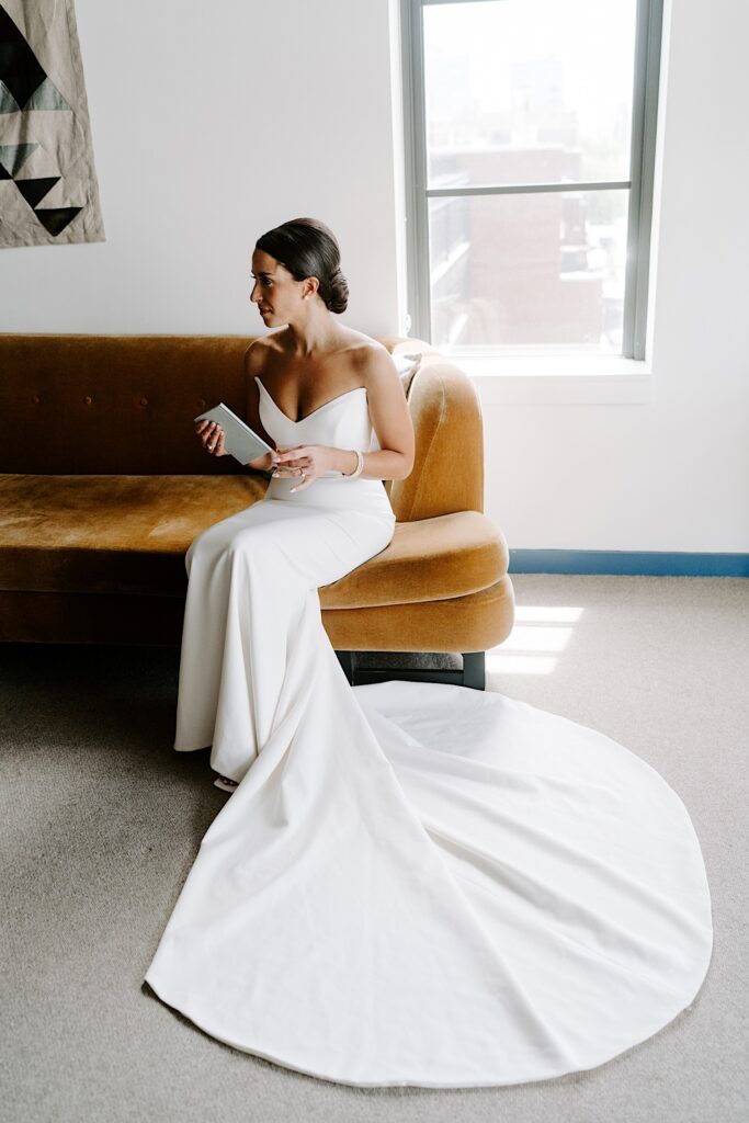 A bride sits on a brown couch in her wedding dress and looks to the left with tears in her eyes after reading her vows