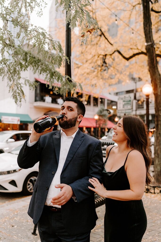 A woman laughs and touches a man's arm as he drinks from a bottle of champagne during their engagement session in Chicago