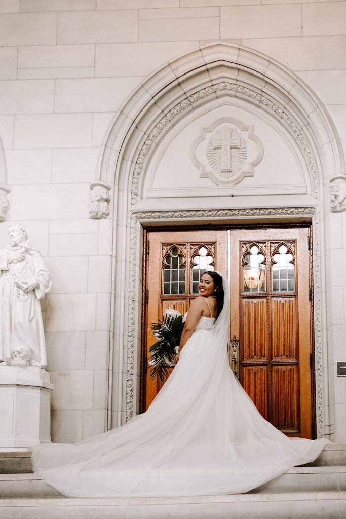 A bride standing at the top of a staircase outside the wooden doors of a church looks over her shoulder and smiles back at the camera