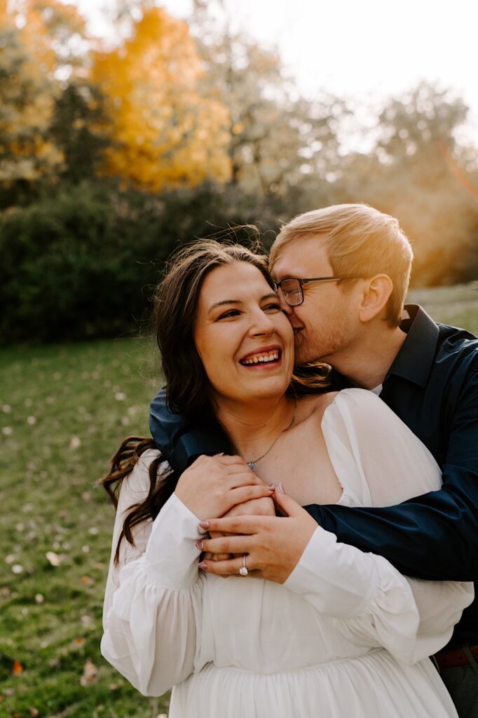 A woman smiles and laughs as a man hugs her from behind and kisses her on the cheek in a forest in the fall