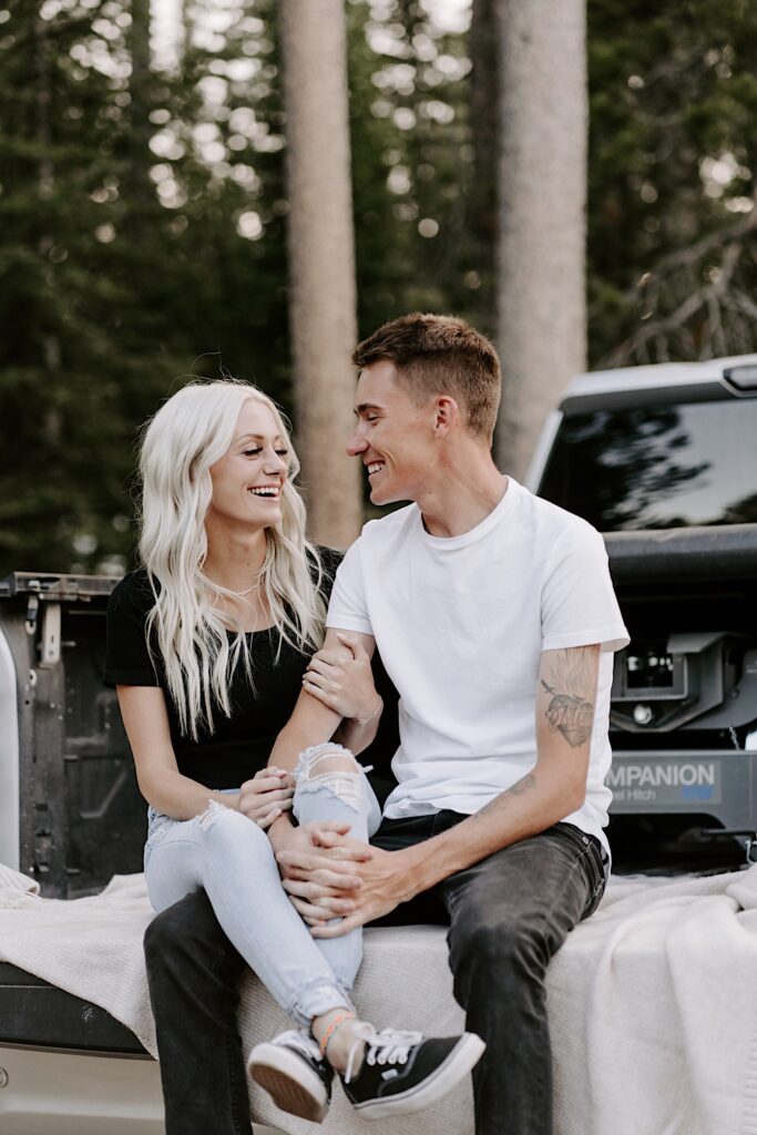 A man and woman sit next to one another in a truck bed and smile at each other in a forest