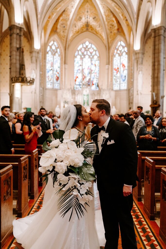 A bride and groom kiss one another in the middle of the aisle as they exit their wedding ceremony in a church as guests one either side of them watch