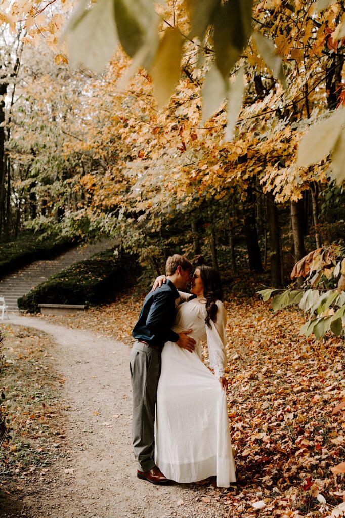 A woman smiles and embraces a man who is going in for a kiss as the two stand on a path covered in leaves during the fall