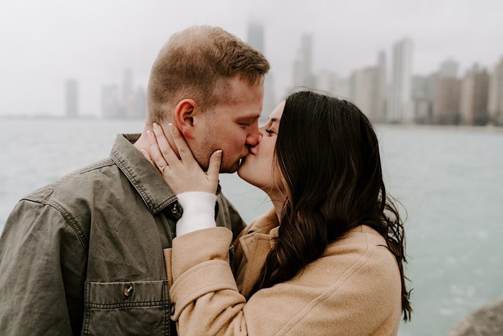 A man and woman kiss in front of Lake Michigan and the Chicago skyline during their Fall Engagement Session at North Avenue Beach