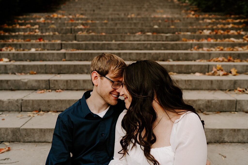 A couple sit on a stone staircase covered in leaves and look at one another while smiling and touching their foreheads together while taking Fall engagement photos in Chicago