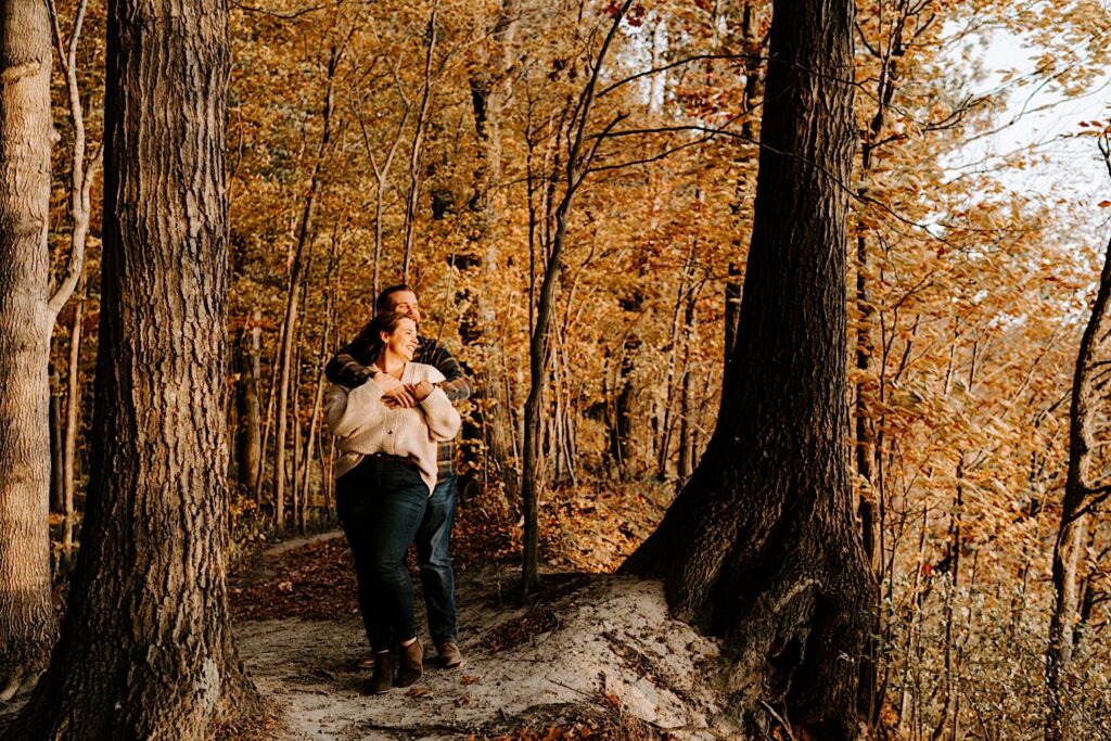 A man stands behind a woman and hugs her as the two look off to the right and smile while surrounded by orange trees during their fall engagement session
