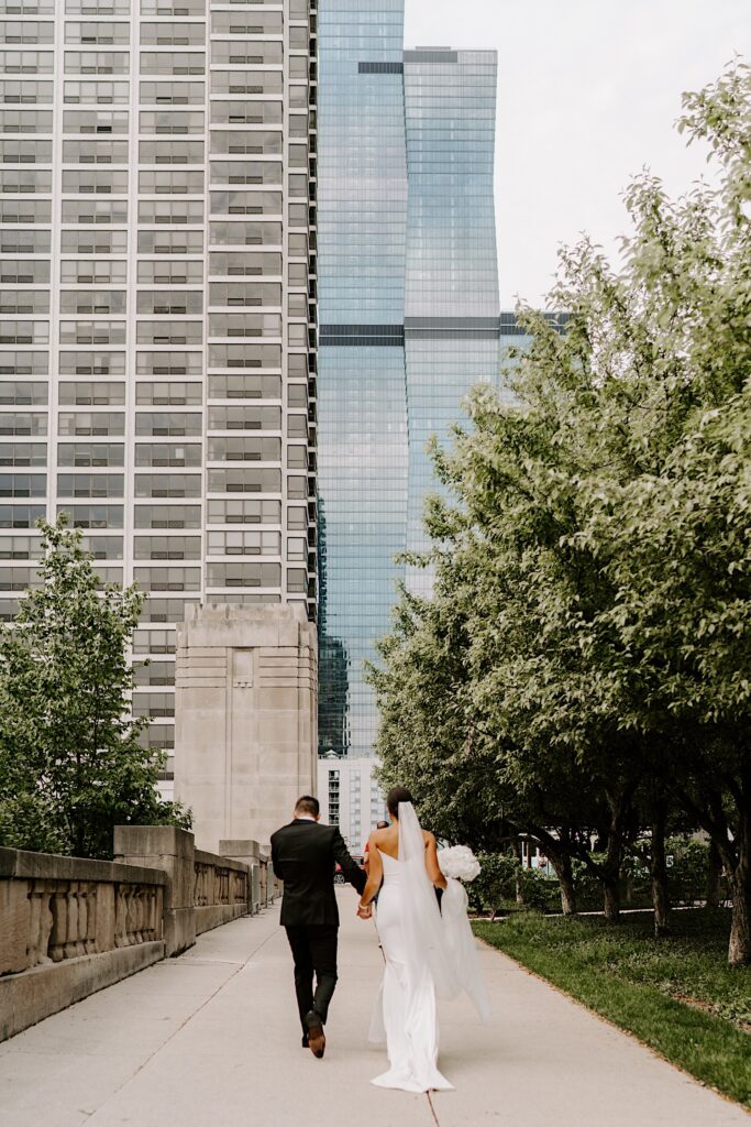A bride and groom walk hand in hand away from the camera towards large buildings in Chicago