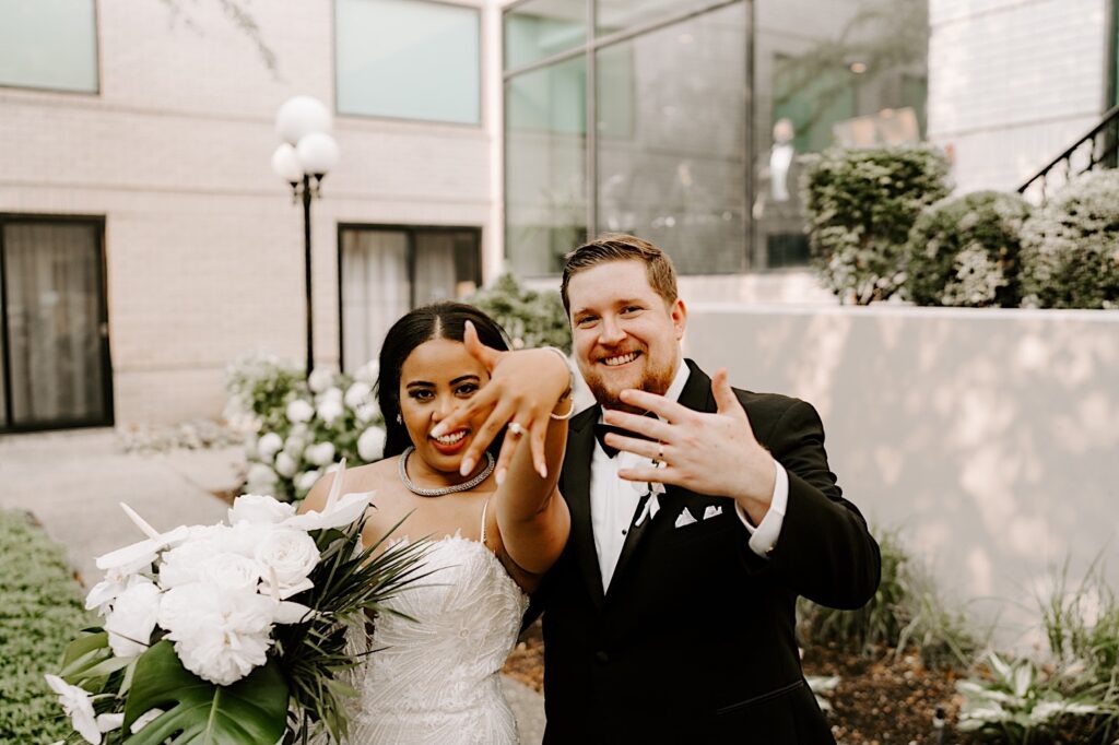 A bride and groom standing outside a building smile as they show their ring hands to the camera