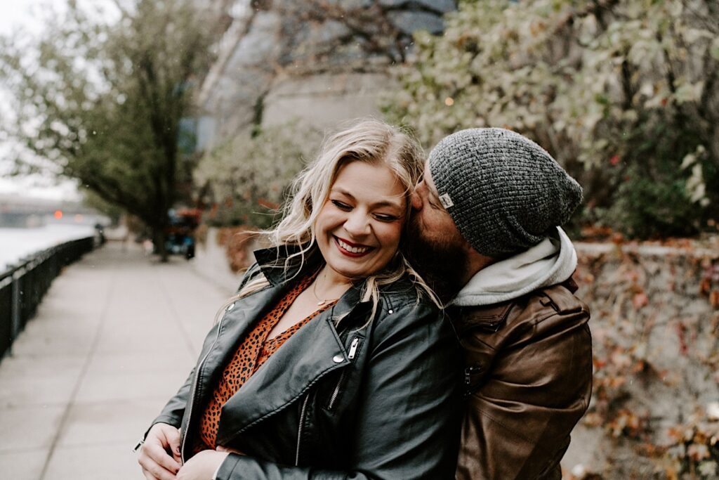 A woman smiles towards the camera with her eyes closed as a man hugs her from behind and kisses her cheek while taking their engagement photos in the fall in Chicago