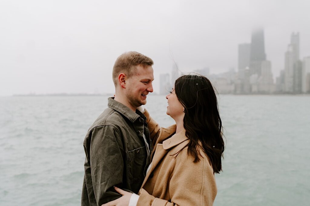 A man and woman embrace and smile at one another while standing in front of Lake Michigan and the Chicago skyline during their engagement session at North Avenue Beach