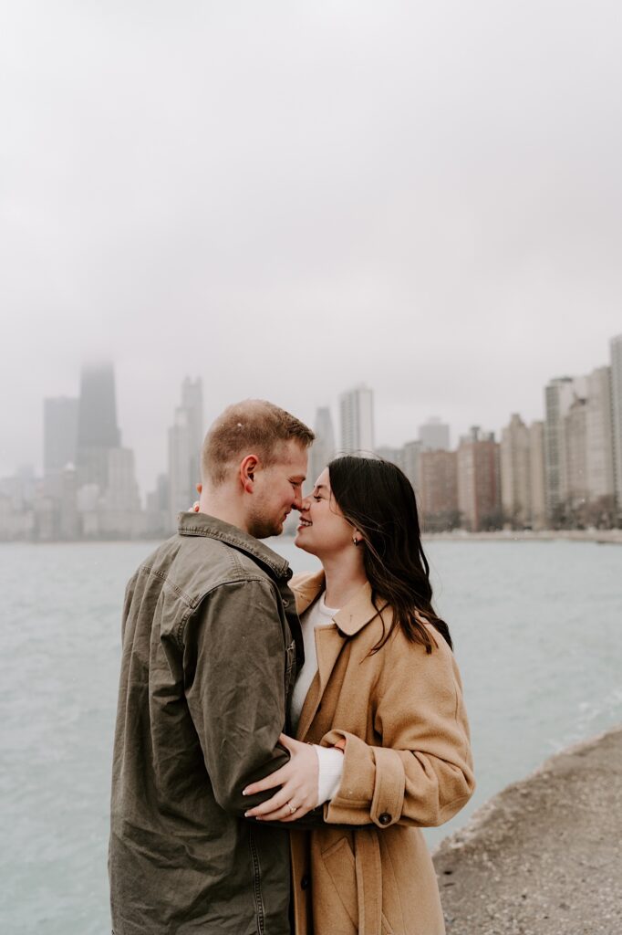 A man and woman embrace each other and smile as they are about to kiss one another with the Chicago skyline and Lake Michigan behind them