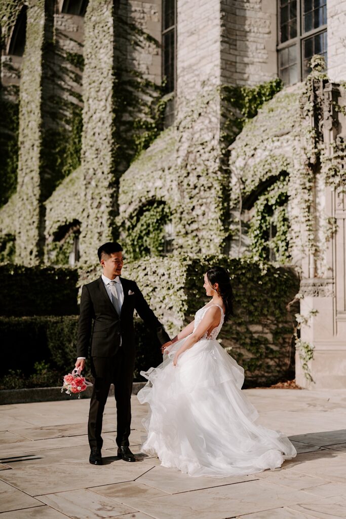 A bride and groom stand outside of a church covered in ivy vines and look at one another while smiling