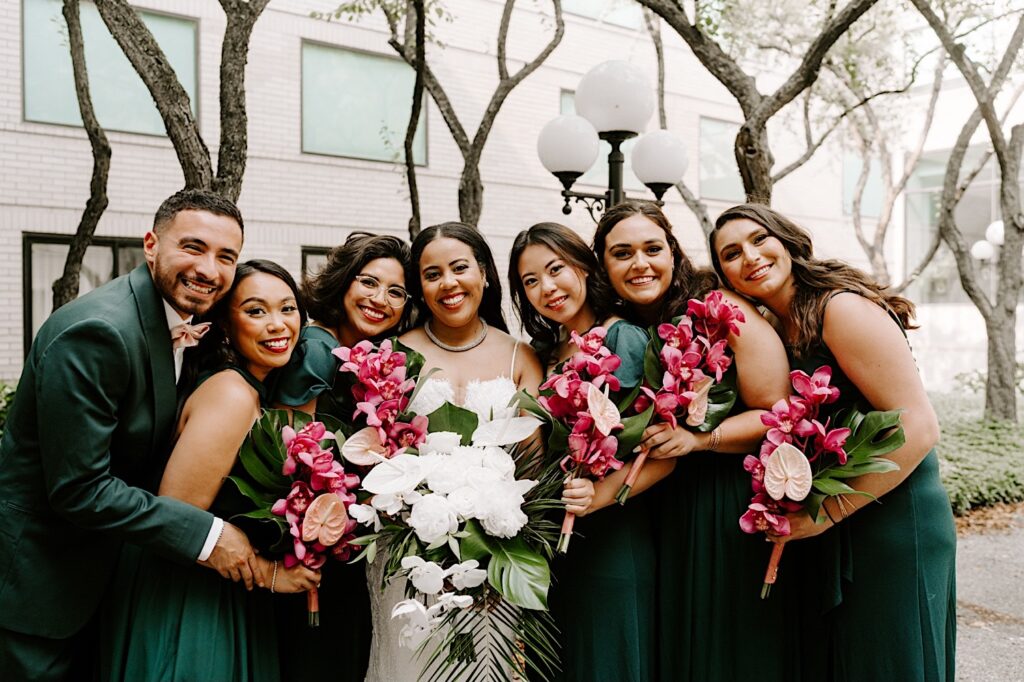 A bride smiles at the camera while standing outside with  her group of 6 wedding party members who are also smiling at the camera