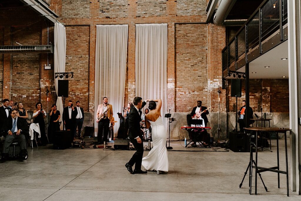 A bride and groom dance in front of a live band inside of a brick building during their wedding reception