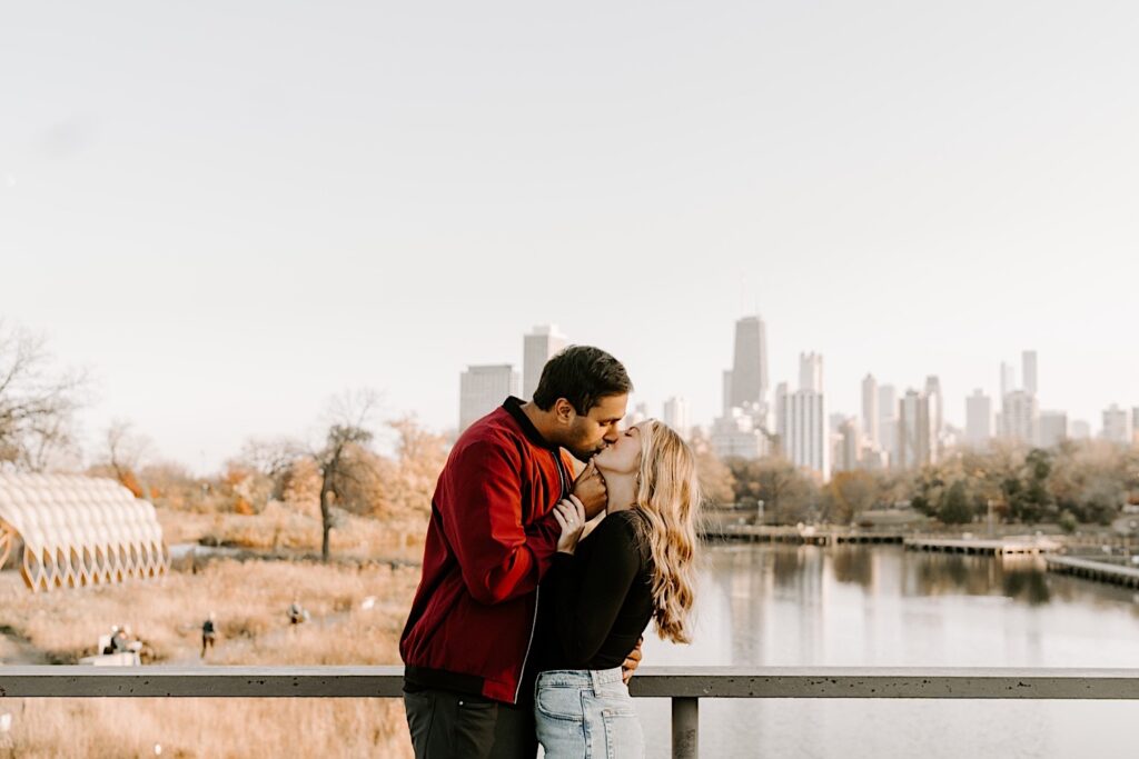 A couple in Chicago's Lincoln Park kiss one another with the skyline and water behind them during their fall engagement photos