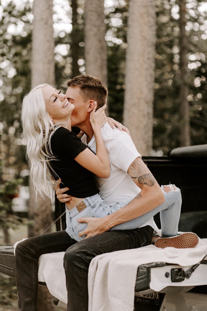 A woman smiles while sitting on a man's lap as he kisses her neck while the two sit on a truck bed in a forest
