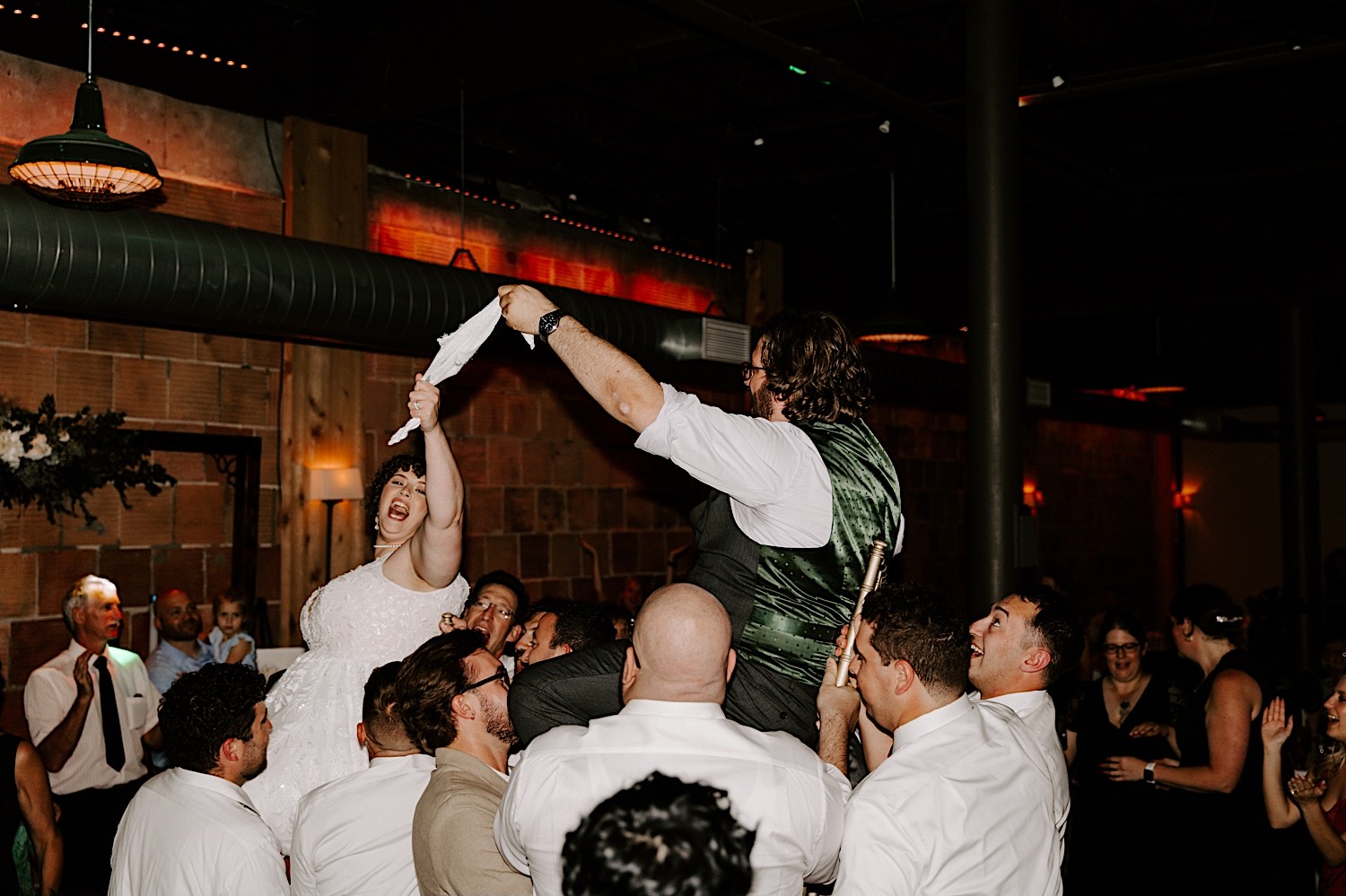 A bride and groom each in chairs are lifted in the air by members of their wedding during their indoor wedding reception, they are each holding a piece of cloth in between them