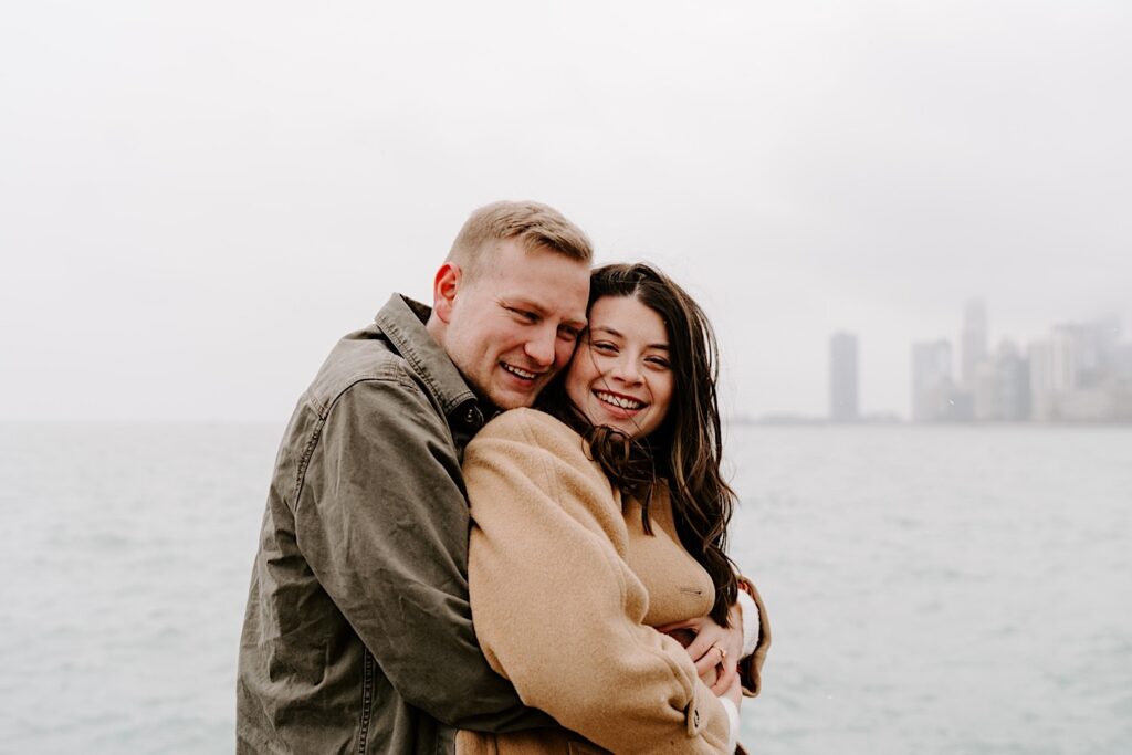During their Fall Engagement Session at North Avenue Beach a man hugs a woman from behind as she smiles at the camera with Lake Michigan and the Chicago skyline behind them