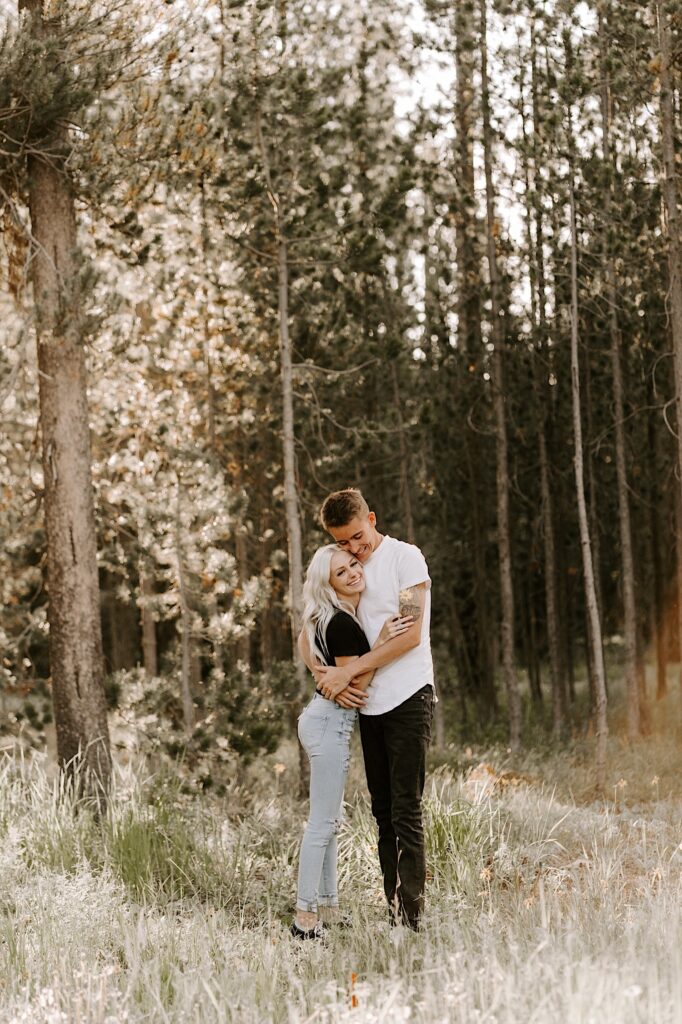 A woman smiles at the camera while being hugged by a man while they stand in the middle of a forest