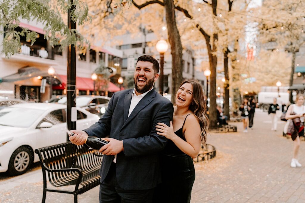 A couple smile while taking their engagement photos in downtown Chicago in the fall, the man is spraying a bottle of champagne that they just opened