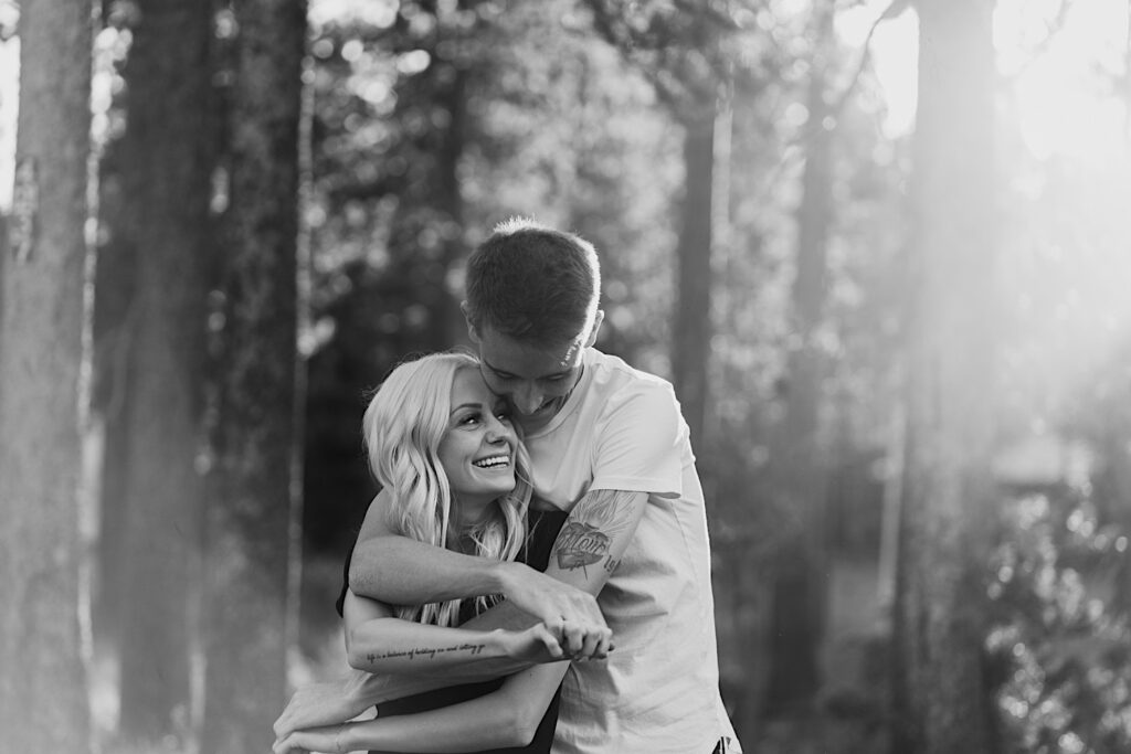 Black and white photo of a woman smiling over her shoulder as a man hugs her from behind in the middle of a forest