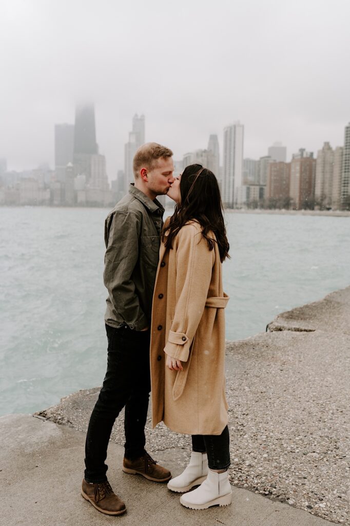 A man and woman kiss one another while standing on the shores of Lake Michigan with the Chicago skyline behind them