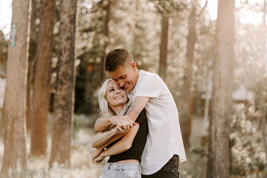 A woman smiles over her shoulder at a man who is hugging her from behind as the two stand in a forest during their engagement session in Busse Woods Forest Preserve