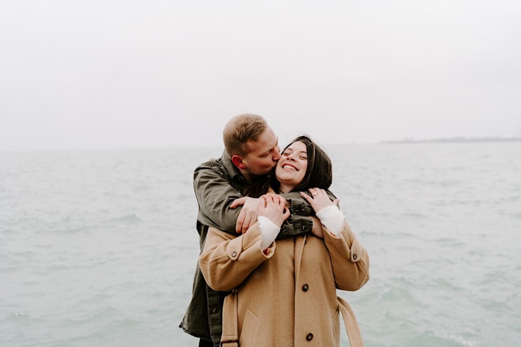 A woman smiles as a man hugs her from behind and kisses her on the cheek while they stand in front of Lake Michigan during their Fall Engagement Session at North Avenue Beach