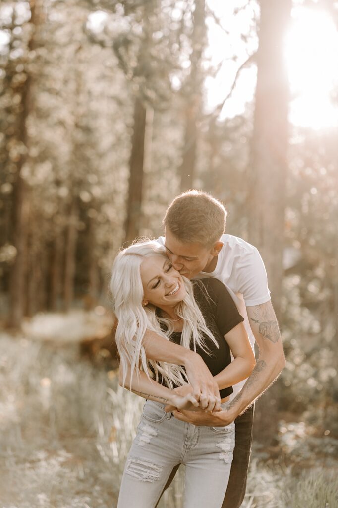 A woman smiles as she is hugged from behind and kissed on the cheek by a man in the middle of a forest