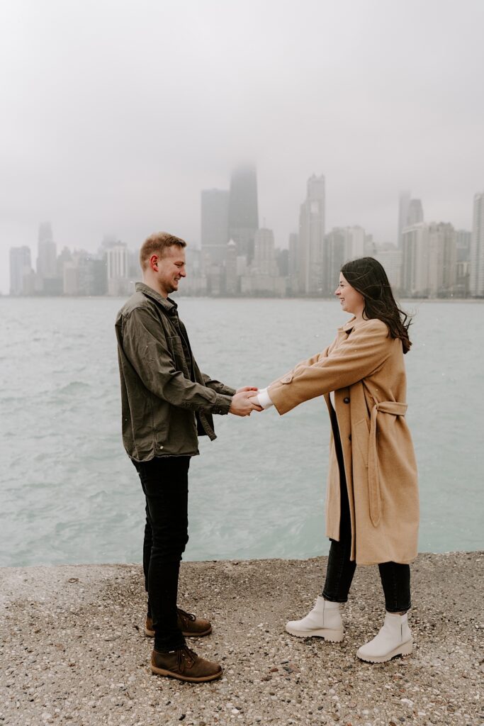 A man and woman face one another and hold hands while smiling at one another standing in front of Lake Michigan and the Chicago skyline