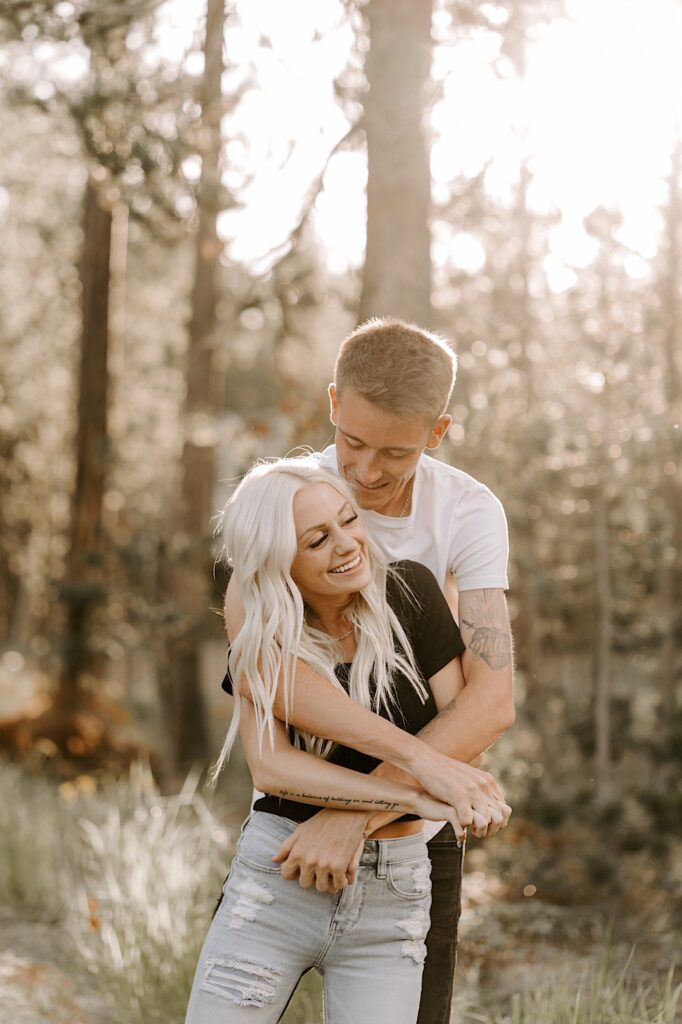 A woman smiles as a man hugs her from behind and smiles at her while they stand in the middle of a forest